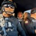 Two Ecuadoran police officers in dark sunglasses, helmets and balaclavas - which entirely obscure their faces - sit in a car in front of the BBC's Orla Guerin, wearing a flak jacket with the word "PRESS" on it.