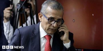 Ollanta Humala speaks on the phone as he leaves court in 2025. He wears a navy suit and red tie
