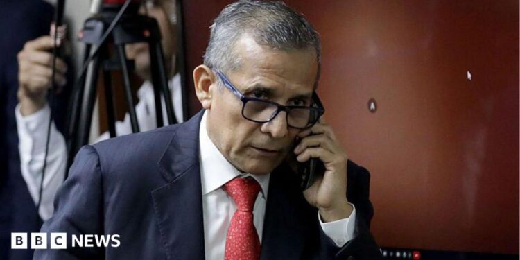 Ollanta Humala speaks on the phone as he leaves court in 2025. He wears a navy suit and red tie