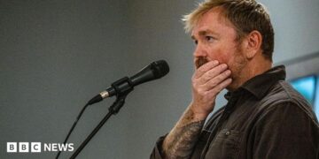 Graham Platner seen on stage at a microphone. his hand is over his mouth
