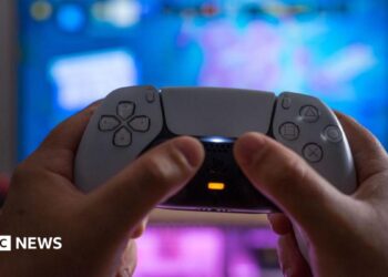 A white PlayStation 5 controller being held up by someone's hands against a blurred TV screen behind it