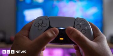 A white PlayStation 5 controller being held up by someone's hands against a blurred TV screen behind it
