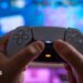 A white PlayStation 5 controller being held up by someone's hands against a blurred TV screen behind it