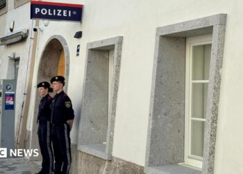 Two police officers stand outside a white-washed building with a police sign in German on the wall