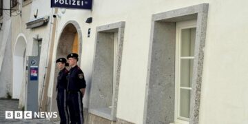Two police officers stand outside a white-washed building with a police sign in German on the wall