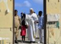 Pope Leo holds hands with a girl wearing a red dress and a man in blue shirt and white trousers. A women in a beige outfit stands alongside them at a doorway. Behind them are blue skies and men dressed in black