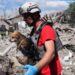 A rescue worker wearing a helmet carries a small dog next to the rubble of a building destroyed by an attack.
