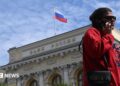 A woman wearing a red jumper and sunglasses, as well as a little bag on her right shoulder, talks on the phone in front of an exterior view of the Central Bank of the Russian Federation building on top of which the Russian flag can be seen