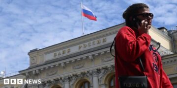 A woman wearing a red jumper and sunglasses, as well as a little bag on her right shoulder, talks on the phone in front of an exterior view of the Central Bank of the Russian Federation building on top of which the Russian flag can be seen