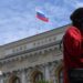 A woman wearing a red jumper and sunglasses, as well as a little bag on her right shoulder, talks on the phone in front of an exterior view of the Central Bank of the Russian Federation building on top of which the Russian flag can be seen
