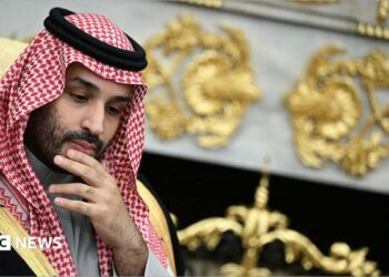 He is rubbing his chin, thinking. He is wearing traditional Arab clothing, with the red and white head dress. He is wearing a black robe with gold edges.