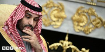 He is rubbing his chin, thinking. He is wearing traditional Arab clothing, with the red and white head dress. He is wearing a black robe with gold edges.