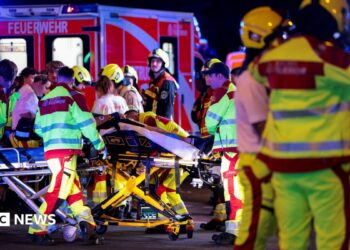 A number of emergency services personnel standing near an ambulance, with a person on a stretcher