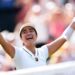 Filipina tennis player Alexandra Eala raises his arms after a Wimbledon match, net in front of her and a blurred image of the crowd in the background.