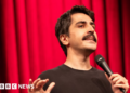 A head-shot posted by a Turkish stand-up comic showing him holding a microphone and standing in front of a red curtain