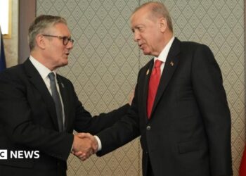 Sir Keir and Erdogan, both wearing dark suits, shake hands. Behind them are the Union Jack and Turkish national flag.