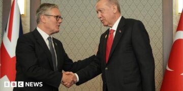 Sir Keir and Erdogan, both wearing dark suits, shake hands. Behind them are the Union Jack and Turkish national flag.