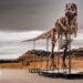 'Gus' the T. rex - an assembled skeleton of a T-Rex dinosaur standing on plinths - stands outside in the Badlands territory of South Dakota on a cloudy grey day against the backdrop of mountains and brown shrubland.