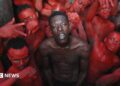 A topless Empereur stands in the centre of a group of young men, who are painted in red.