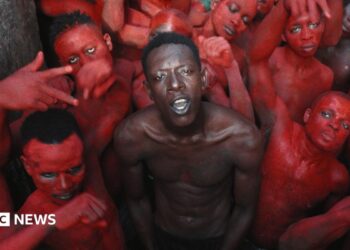 A topless Empereur stands in the centre of a group of young men, who are painted in red.