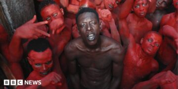 A topless Empereur stands in the centre of a group of young men, who are painted in red.