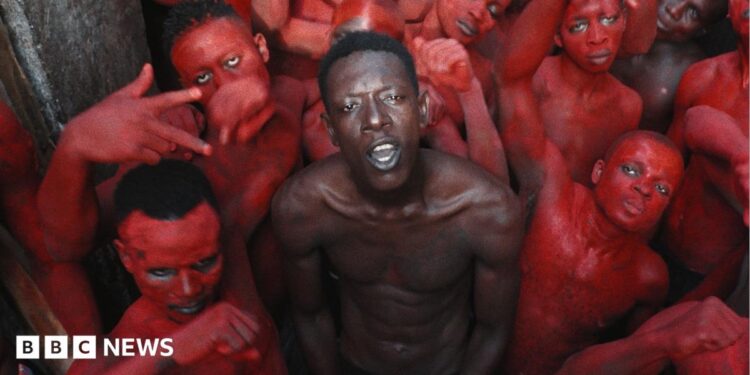 A topless Empereur stands in the centre of a group of young men, who are painted in red.