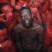 A topless Empereur stands in the centre of a group of young men, who are painted in red.