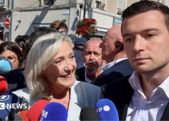 A woman stands in a crowd smiling in front of microphones while a man to her right stands beside her with no expression