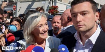 A woman stands in a crowd smiling in front of microphones while a man to her right stands beside her with no expression