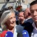 A woman stands in a crowd smiling in front of microphones while a man to her right stands beside her with no expression