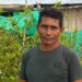 Coca farmer Perea standing among his plants. Behind him are green sheets hanging up in front of a building.