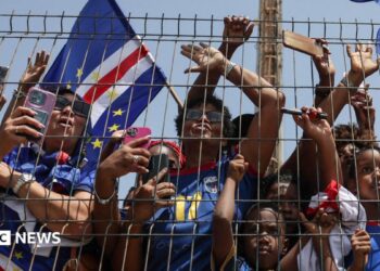 Fans wearing the Cape Verde kit press against a metal fence.