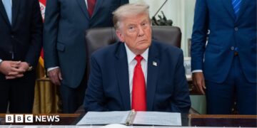 US President Donald Trump sitting in the White House wearing a blue suit jacket, white shirt and red tie.