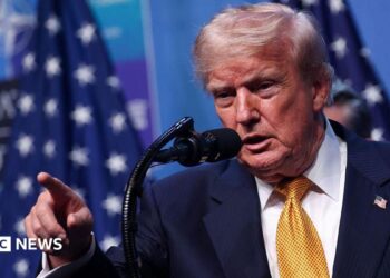 US President Donald Trump wearing a yellow tie, white shirt, blue suit and American flag pin. He stands at a microphone and is pointing, his mouth open.