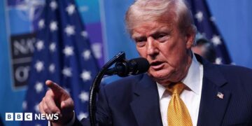 US President Donald Trump wearing a yellow tie, white shirt, blue suit and American flag pin. He stands at a microphone and is pointing, his mouth open.