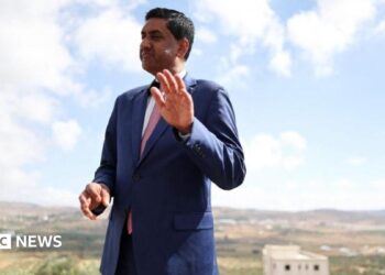 Khanna, who is dressed in a blue suit, white shirt and pink tie, raises his hand as he is photographed outdoors with a rural town landscape behind him