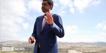 Khanna, who is dressed in a blue suit, white shirt and pink tie, raises his hand as he is photographed outdoors with a rural town landscape behind him
