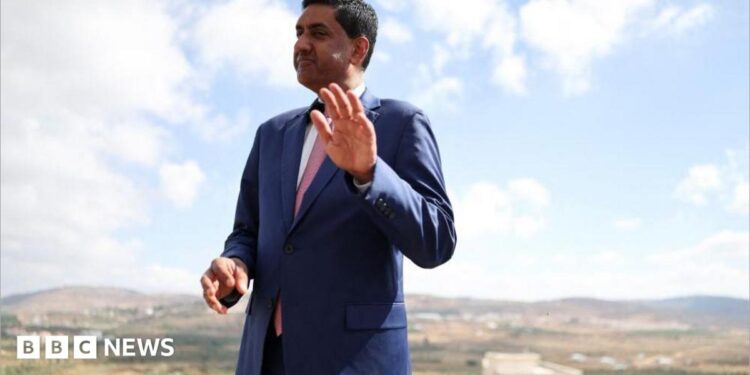 Khanna, who is dressed in a blue suit, white shirt and pink tie, raises his hand as he is photographed outdoors with a rural town landscape behind him