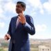 Khanna, who is dressed in a blue suit, white shirt and pink tie, raises his hand as he is photographed outdoors with a rural town landscape behind him