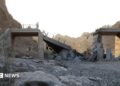 A damaged bridge with people standing on the remaining structure with mountains in the background, and rubble and rocks in the foreground.