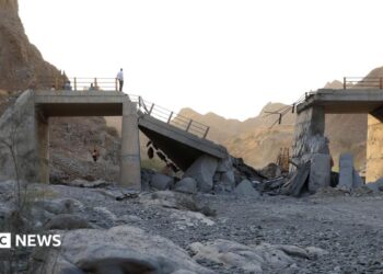 A damaged bridge with people standing on the remaining structure with mountains in the background, and rubble and rocks in the foreground.
