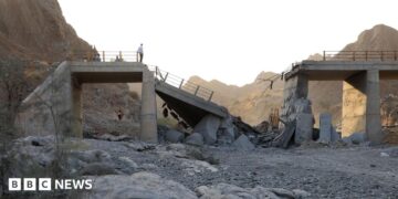 A damaged bridge with people standing on the remaining structure with mountains in the background, and rubble and rocks in the foreground.