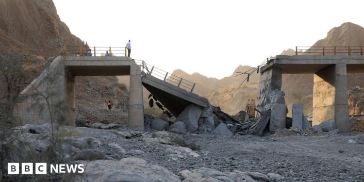 A damaged bridge with people standing on the remaining structure with mountains in the background, and rubble and rocks in the foreground.