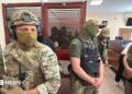 Men in military fatigues stand in a courtroom with a person sitting in a glass room