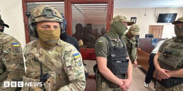 Men in military fatigues stand in a courtroom with a person sitting in a glass room