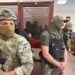 Men in military fatigues stand in a courtroom with a person sitting in a glass room