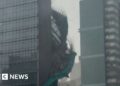 Bamboo and green plastic sheeting are seen falling down the side of a glass skyscraper.