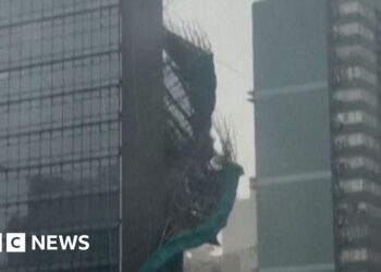 Bamboo and green plastic sheeting are seen falling down the side of a glass skyscraper.