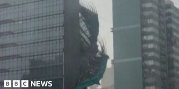 Bamboo and green plastic sheeting are seen falling down the side of a glass skyscraper.
