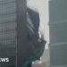 Bamboo and green plastic sheeting are seen falling down the side of a glass skyscraper.